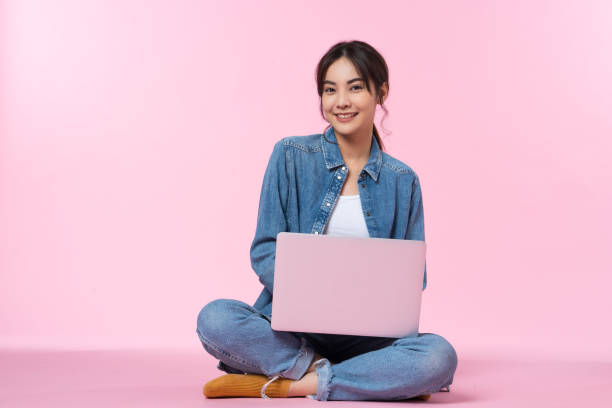 Young Asian college student sits cross-legged on floor working with a laptop computer, looking directly at the camera in a studio shot on a pink background