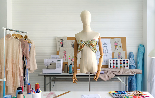 Mannequin standing with colorful fabrics in fashion designer room. Sewing machine and various sewing related items on the table.
