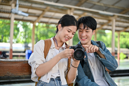 Cute and lovely young Asian couple is talking and looking at their picture on a camera together while waiting for their train at the railway station.