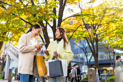 Happy Asian woman friends using navigator app on mobile phone during shopping together at Shibuya district, Tokyo, Japan. Attractive girl enjoy outdoor lifestyle travel city street on holiday vacation.