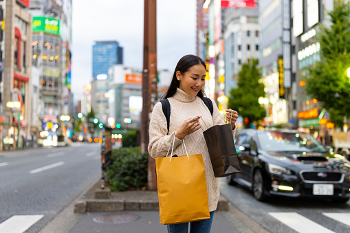Happy Asian woman holding shopping bag walking and shopping at Shibuya district, Tokyo, Japan in evening. Attractive girl enjoy outdoor lifestyle travel urban city street in autumn holiday vacation.