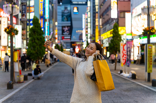 Happy Asian woman using mobile phone taking selfie during shopping at Tokyo city, Japan. Attractive girl enjoy outdoor lifestyle travel city street with using wireless technology on holiday vacation.
