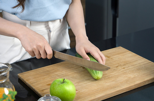 Closeup woman hand holding knife cutting green cucumber on a wooden chop board.