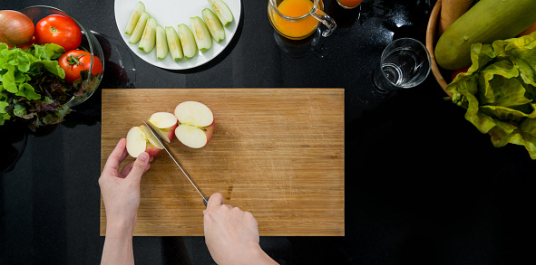 Closeup hand holding knife cutting red apple on a wooden chop board (Top View). Sliced fruits are placed on a plate. The kitchen counter full of various kinds of vegetables and fruit juice.