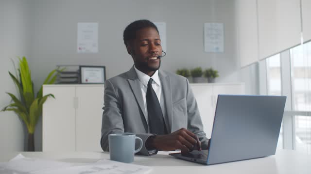 Happy black businessman with headset having online conference on laptop in office