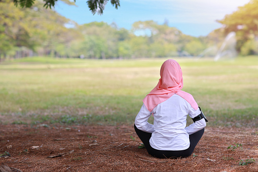 Rear view young asian muslim woman sitting on grass, enjoying meditation. Peaceful girl in sportswear with pink hijab practice yoga in nature green trees with peace and calm. Healthy and sport concept