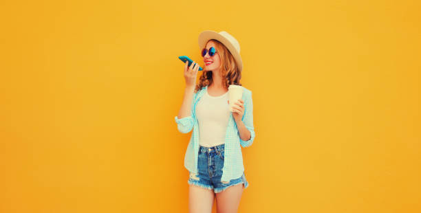 istock Happy young woman using voice command recorder, assistant or takes calling wearing straw hat, shorts on orange background 1497097414