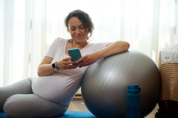 Smiling pregnant woman leaning on a fit ball, relaxing with smartphone on exercise mat after prenatal fitness at home.