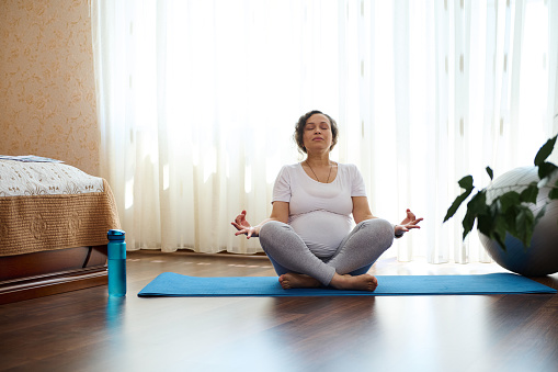 Spiritual and emotional concept of harmony in pregnancy and maternity time. Pregnant woman practice yoga, sitting on fitness mat on the floor at home, reaching zen, balance and harmony. Body conscious