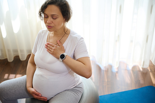 Beautiful serene pregnant adult woman sitting on fitness ball, doing meditation and pranayama breathing exercises for healthy pregnancy, preparing body for childbirth, putting hands of chest and belly