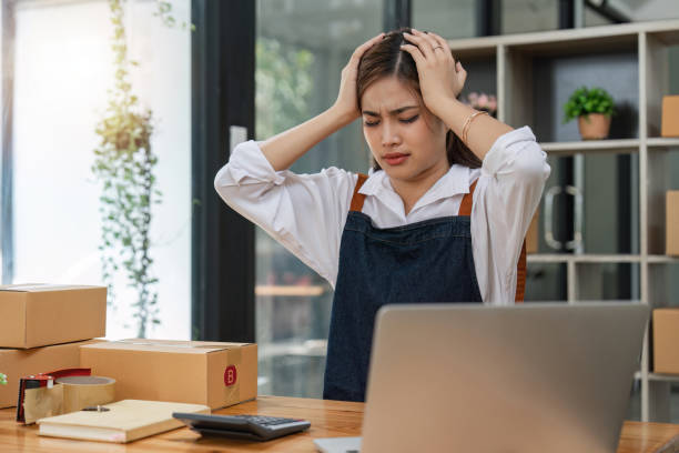 Small online business owner Asian woman wearing apron stressed headache taking notes with parcel box at home Small online business owner Asian woman wearing apron stressed headache taking notes with parcel box at home. newspaper seller stock pictures, royalty-free photos & images