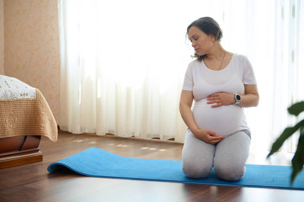 Pretty pregnant woman in late pregnancy, strokes belly, sitting in hero pose on yoga mat at home, dreamily looking aside