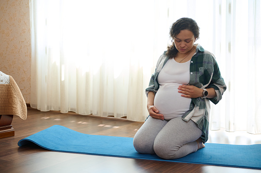 Beautiful multi ethnic adult pregnant woman, sitting hero in pose on a blue fitness mat, holding her belly, during relaxation and breathing exercises in cozy home interior, against window background.