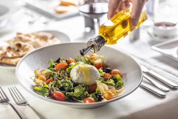 A hand of a person pours olive oil on top of Buffalo mozzarella ball on top of a healthy Italian salad. stock photo