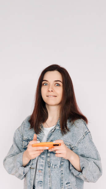 portrait of a young woman in a casual denim jacket with a smartphone playing a game, isolated against a gray background, conveying indecision and insecurity - bolero jacket imagens e fotografias de stock