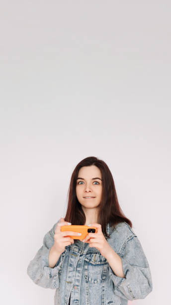 indecisive and insecure: young woman in denim jacket playing smartphone game, isolated on gray background. emphasizing uncertainty and doubt - bolero jacket imagens e fotografias de stock