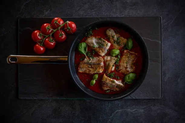 Delicious and healthy white fish dish with spicy tomato sauce. Served hot and ready to eat in a frying pan isolated on dark background from above. Low Lighting