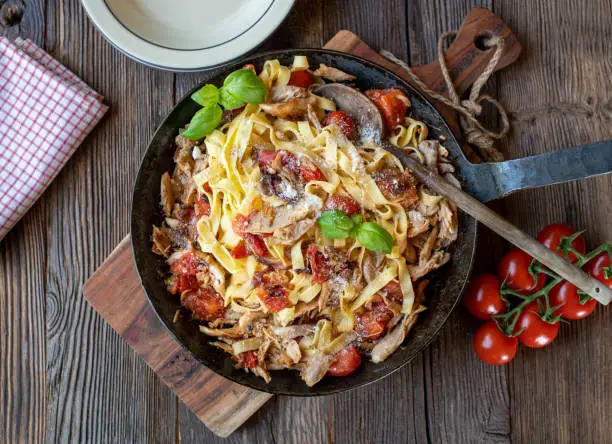 Delicious mediterranean pasta dish with chicken meat, cherry tomatoes, red onions, garlic and herbs. Cooked tagliatelle noodles. Served in a rustic iron pan on wooden table background. Top view