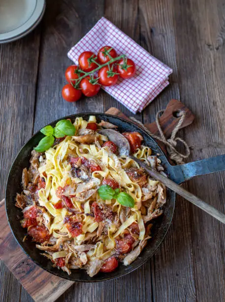 Delicious homemade italian pasta dish with roasted chicken meat, tagliatelle noodles, tomatoes, onions, garlic, herbs and grated parmesan cheese in a rustic frying pan with basil on wooden table from above.
