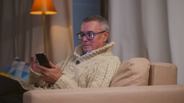 Senior man using smartphone browsing internet sitting on couch at home.