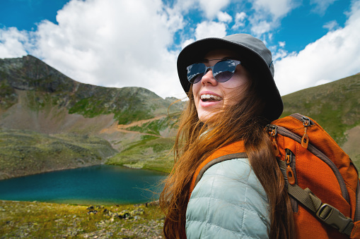 portrait of a girl in the mountains standing looking at the mountain landscape. Traveler portrait. The concept of adventure, travel and hiking. Happy woman in a cap enjoys the sunshine while hiking in the mountains. Tourist with glasses.