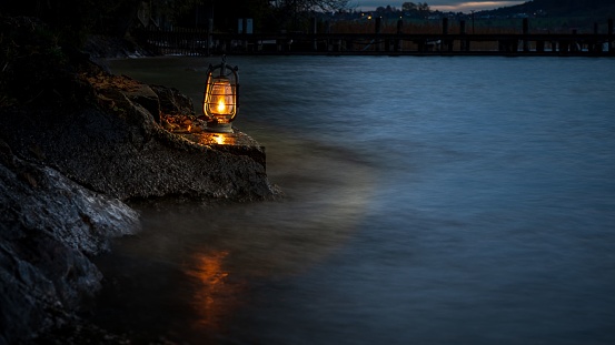 A lit lantern rests on the rock in the dark near tranquil waters of a beach during the night