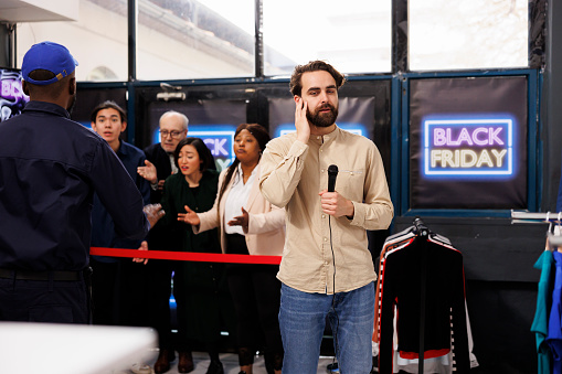 Reporter broadcasting about Black Friday madness from shopping mall. Young man TV journalist holding microphone making live reportage while standing against angry mad shoppers waiting for sales