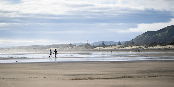 Couple holding hands and walking barefoot on Muriwai beach. Unrecognizable people walking in the distance. Auckland.