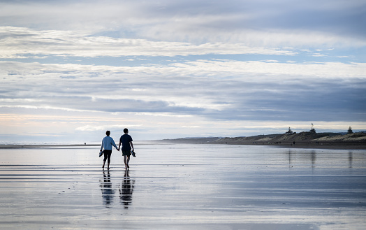 Couple holding hands, holding shoes, and walking barefoot on Muriwai beach. Auckland.