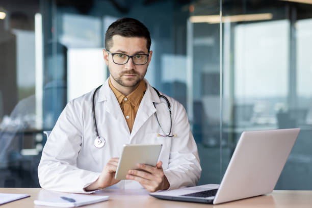 portrait d’un jeune médecin spécialiste assis dans le bureau à la table en blouse blanche et avec un stéthoscope et regardant la caméra - service computer training office photos et images de collection