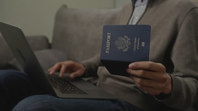 Unrecognised Man Holding US Passport and Typing on Laptop. Person filling form with passport ID number.