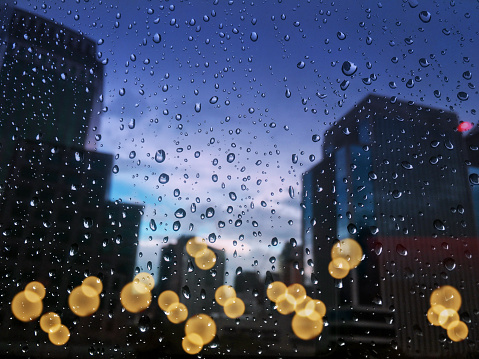 Raindrops on glass with cityscape in the background blurred and the background city bokeh light. City life in the rainy season evening, abstract urban background.