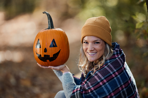 Portrait of happy woman holding Jack O' Lantern in autumn day at the park and looking at camera.