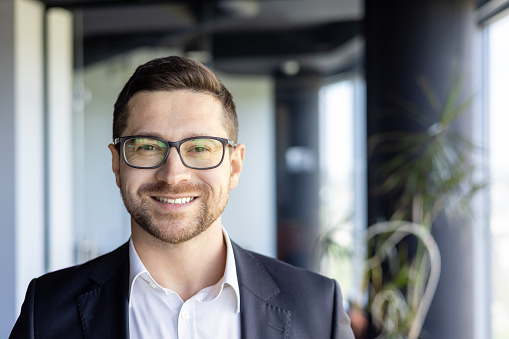 Close-up photo portrait of young successful entrepreneur, businessman investor wearing glasses at workplace smiling and looking at camera.