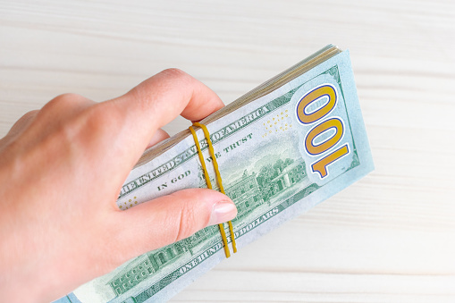 A stack of one hundred dollar bills is held by a woman's hand. A stack of one hundred dollar bills tied with a rubber band in a hand on a light background.
