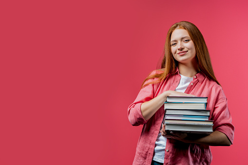 Clever student holds stack of university books from college library on pink background. Happy red-haired girl smiles, she is happy to graduate. Copy space. High quality photo
