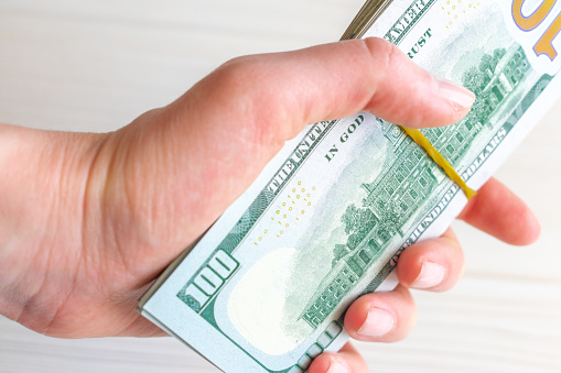 A stack of one hundred dollar bills is held by a woman's hand. A stack of one hundred dollar bills tied with a rubber band in a hand on a light background.