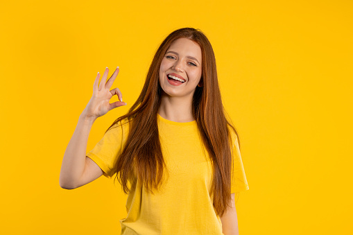Pretty woman makes hand sign okay, ok gesture. Happy ginger girl, correct perfect choice, great deal, yellow background. Positive female model smiles to camera, approval, trust concept. quality photo