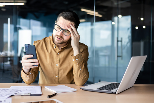Worried young male businessman received bad news on phone, sitting in office at desk and upset reading message, mail, holding head, thinking about problem solution.