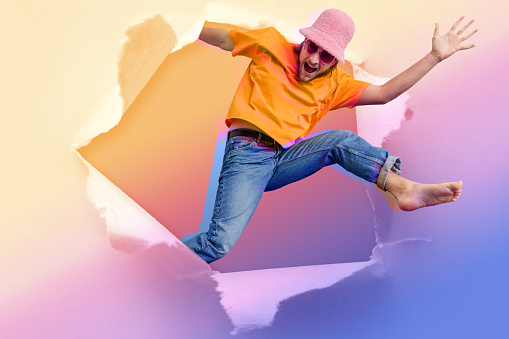 A digitally manipulated image of a young man wearing a bucket hat and sunglasses jumping and leaping through a ripped hole in a piece of paper while looking at the camera and looking excited.