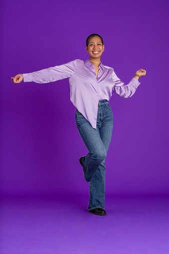 A studio portrait shot of a young woman wearing a silky purple blouse, dancing in front of a purple backdrop while looking at the camera and smiling with her arms out as she dances.