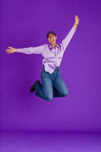 A studio portrait shot of a young woman wearing a silky purple blouse and baseball cap, jumping in front of a purple backdrop while looking at the camera and smiling with her arms in the air.