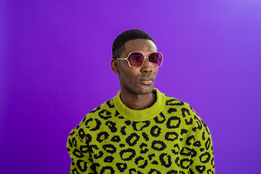A studio portrait shot of a young man wearing a leopard print jumper and sunglasses, standing in front of a purple backdrop and looking off to the side with a contented look on his face.