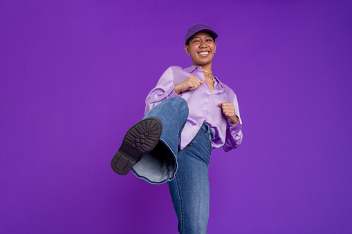 A studio portrait shot of a young woman wearing a silky purple blouse and baseball cap, standing in front of a purple backdrop while looking at the camera and smiling. She is in a kicking stance with one foot in the air and fists clenched at the ready.