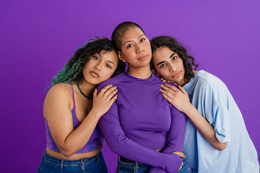 A studio portrait shot of a two female friends and their male friend, standing together in front of a purple backdrop. They are embracing each other while looking at the camera with a contented look on their faces and they are resting their heads on each others.
