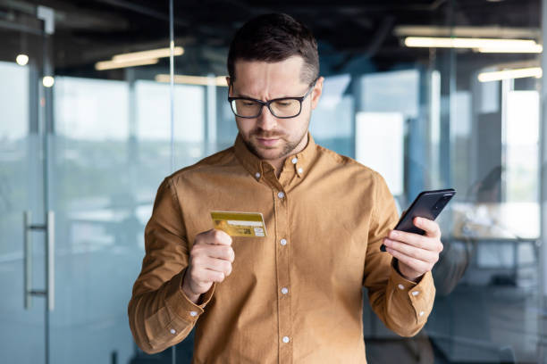 Pensive young man in glasses standing in office with phone and worriedly looking at credit card. Problem with transfer, payment for online shopping, fraud, bankruptcy Pensive young man in glasses standing in office with phone and worriedly looking at credit card. Problem with transfer, payment for online shopping, fraud, bankruptcy. prevention stock pictures, royalty-free photos & images