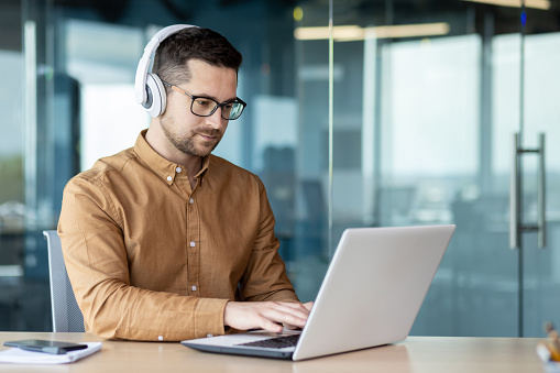 A young man sits at a workplace in the office, works, studies on a laptop with headphones. Listens to the webinar, music, course.
