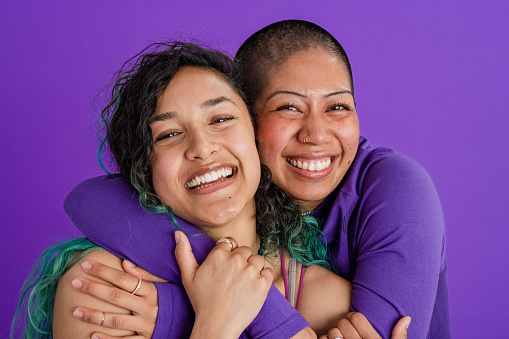A studio portrait shot of a two female friends, standing together in front of a purple backdrop. They are embracing each other while looking at the camera and smiling.