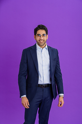 A studio portrait shot of a mid adult man wearing a suit and eyeglasses, standing in front of a purple backdrop while looking at the camera and smiling.