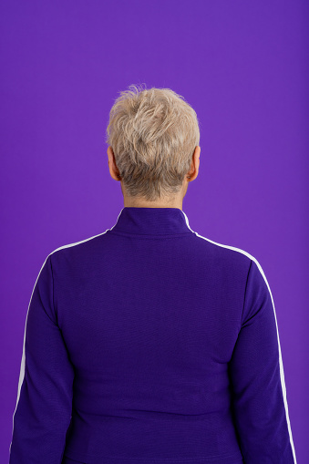 A rear view studio portrait shot of a mature woman wearing a sporty jacket, standing in front of a purple backdrop with her back to the camera.